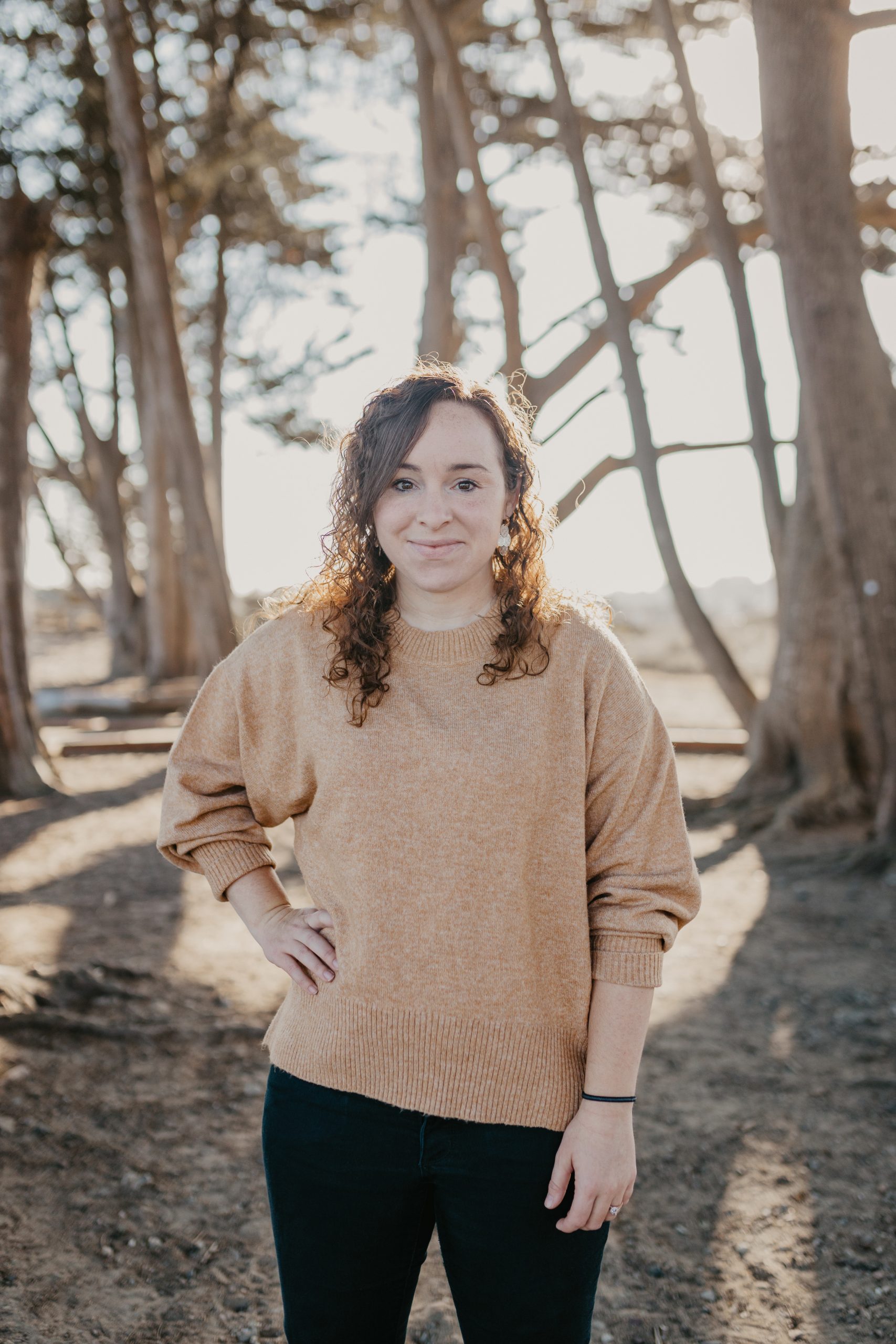 Jen Edwell Woman with brown wavy hair is standing in front of trees with the morning sun behind her. She is wearing a carmel short and smiling.